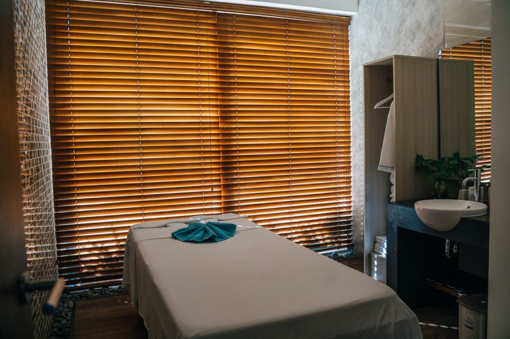 A tranquil massage room with a treatment table covered in a light sheet and a blue towel, backlit by wooden blinds, evok