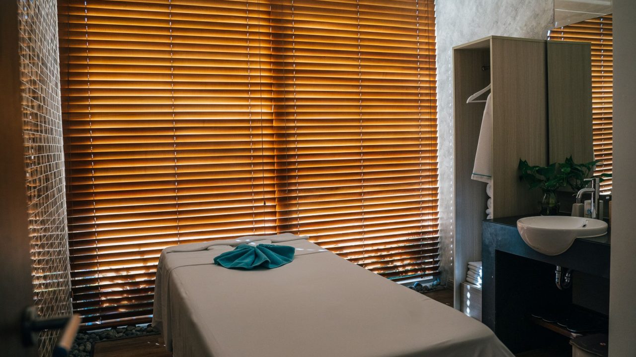 A tranquil massage room with a treatment table covered in a light sheet and a blue towel, backlit by wooden blinds, evok