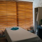 A tranquil massage room with a treatment table covered in a light sheet and a blue towel, backlit by wooden blinds, evok