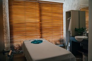 A tranquil massage room with a treatment table covered in a light sheet and a blue towel, backlit by wooden blinds, evok