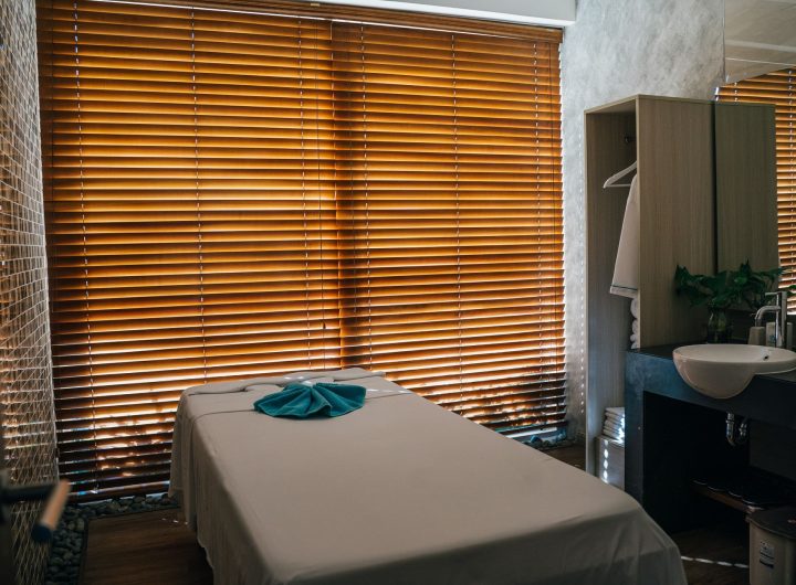 A tranquil massage room with a treatment table covered in a light sheet and a blue towel, backlit by wooden blinds, evok